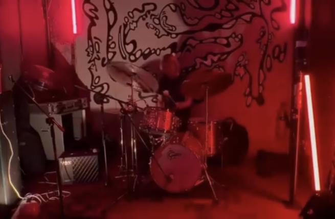 A drum kit in the middle of the room played by Oli Reeves infront of a wall with paintings of distorted faces.