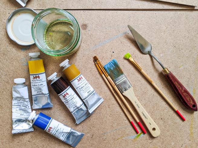 An artist's workspace featuring oil paint tubes, paintbrushes, a palette knife, and a jar of solvent on a wooden surface. The tools are neatly arranged, ready for painting.