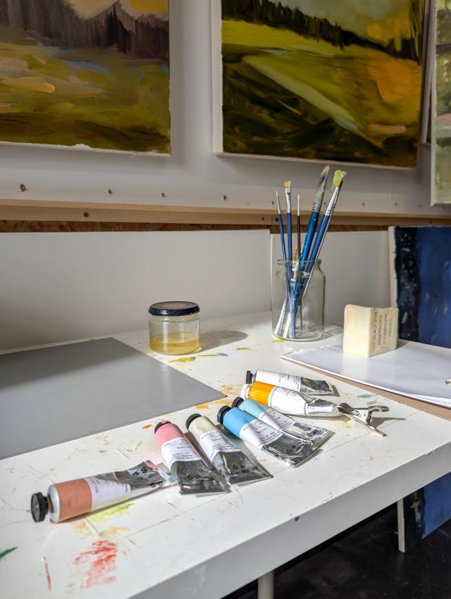 An artist's workspace with paint tubes, brushes in a glass jar, a palette, and a sketchbook on a paint-stained table. Landscape paintings hang on the wall in the background.