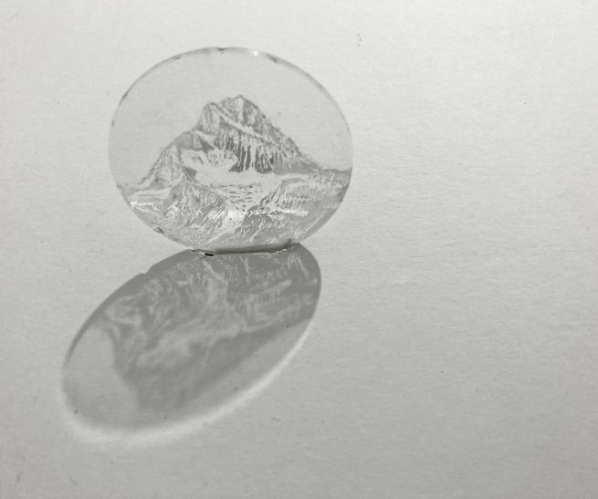 A small circular glass disc etched with a drawing of a mountain on it in black and white. It is attached to a wall and gives a shadow underneath.