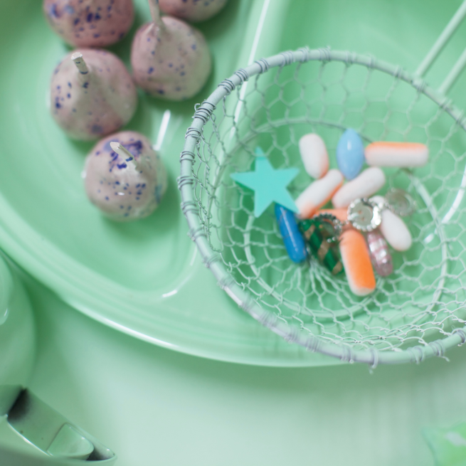 A pastel green hospital food tray on a pastel green table. In the left half of the tray are brown and purple spotted balls on sticks. In the right side of the tray is a wire basket filled with colourful pills.