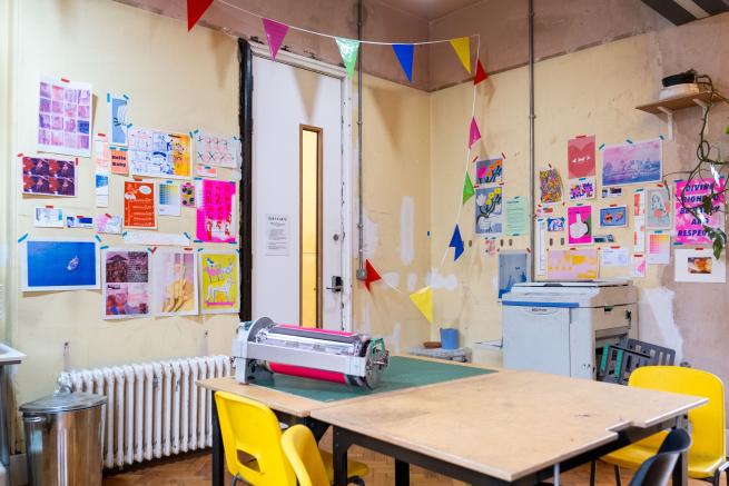an image shows the riso room, with a riso drum on the table, and prints hung on the walls around the room