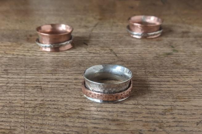 Three rings sit on a wooden surface. One on the foreground is silver with a copper spinning band, the two in the background are copper with a silver 