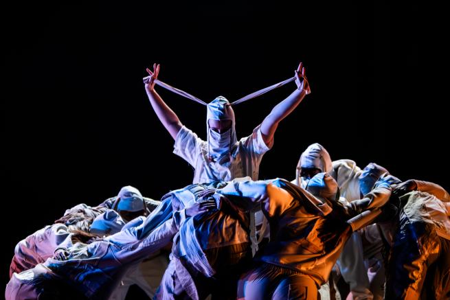 Dancer surrounded by a huddle wears a mask and looks directly at the camera