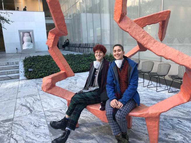 Two people sit on a vibrant orange abstract sculpture in the MoMA sculpture garden, with modern museum architecture and other art installations in the background.