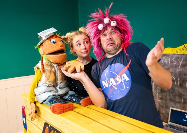 Two actors stand at a piano with a puppet sat on top