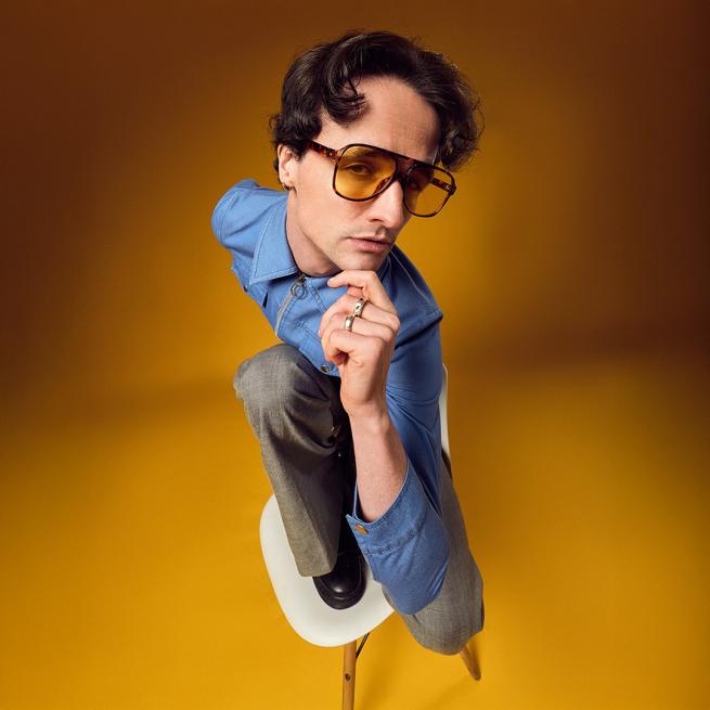 A man in yellow sunglasses and blue shirt poses on a chair and smoulders with two fingers held to his chin.