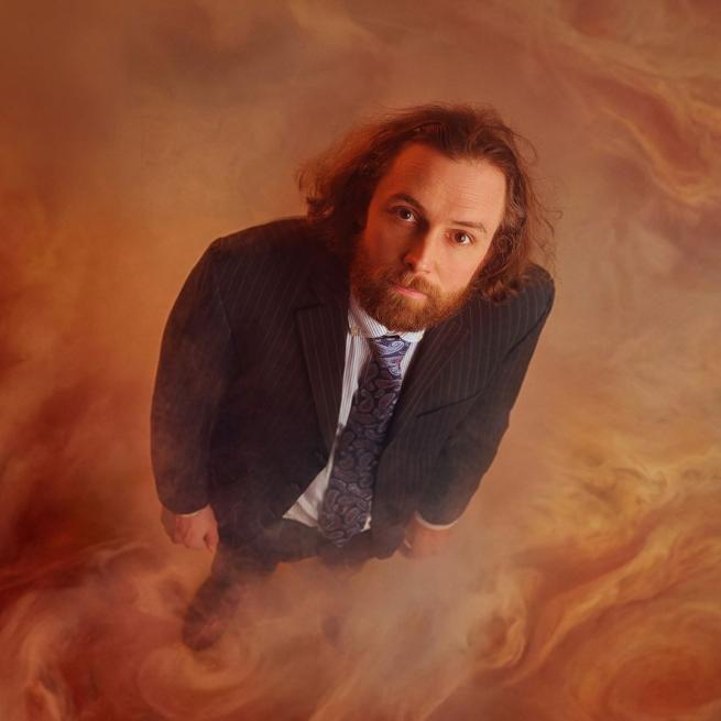 Viewed from above, Rob Auton stands in a suit and looks up at the camera, surrounded by swirling orange smoke.