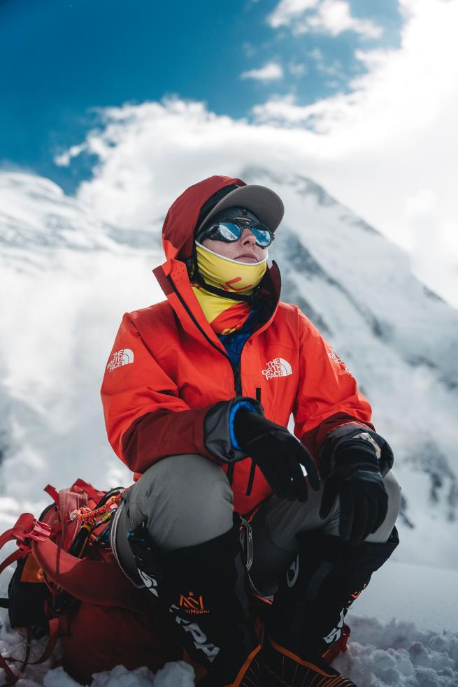 A woman sits at in the snow with a snow-capped mountain behind her, she is in full snow climbing gear.