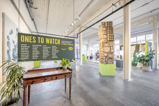 A wide-angle view of the entrance to the Gallery. There is a dark green wooden board suspended over a table, with Ones to Watch and a list of the exhibiting artists printed onto it. Artwork in the exhibition is visible in the background, including a giant tower made from brown packaging materials, positioned on a chartreuse plinth.