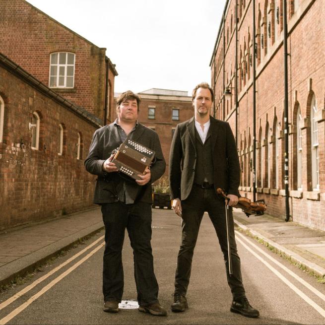 Spiers and Boden stand in the middle of a street of red brick buildings. Both wear casual suit jackets, one holds an accordion and the other a violin.
