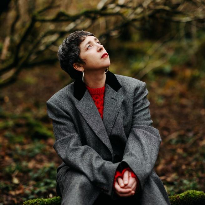 Lisa O'Neill sits in an autumnal forest, wearing a red jumper and grey blazer as she looks skywards.
