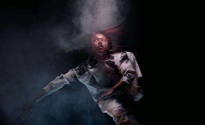 In a dark background, and dancer is captured mid-movement surrounded by a cloud of smoke.