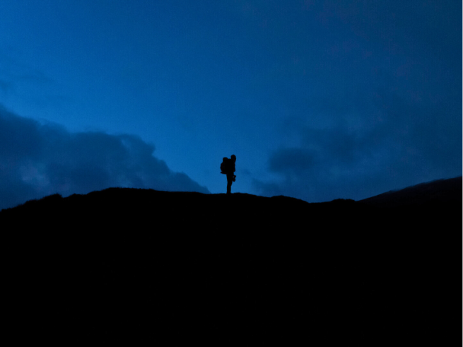 A silhouette of a man stood at the top of a mountain at night time.