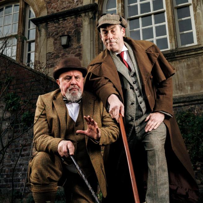 Peter Clifford and the Great Baldini portray a suited Holmes and Watson, who point their walking sticks towards a clue off-camera in the grounds of a grand, old-fashioned building.