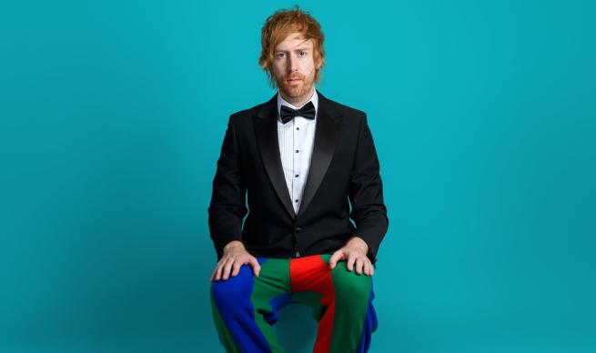 Ginger haired man sat wearing tuxedo jacket, bowtie and multi coloured jesters trousers. He could do with a shave