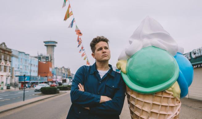 A man stands with arms folded staring thoughtfully into the distance he is leaning against a giant ice cream cone in a seaside resort, the street is festooned with bunting