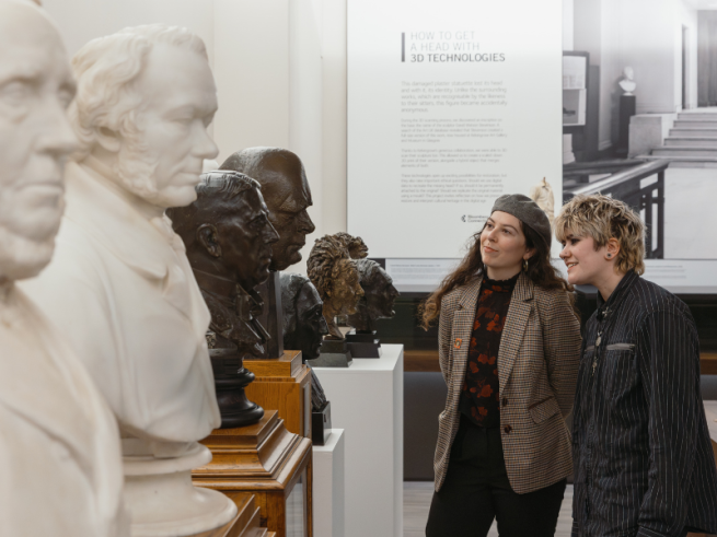 Two people looking at a row of portrait busts made of bronze and marble, stood on plinths