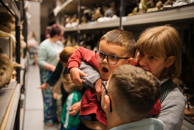 Photograph of an adult picking up a child to look at an object within a museum's storage.
