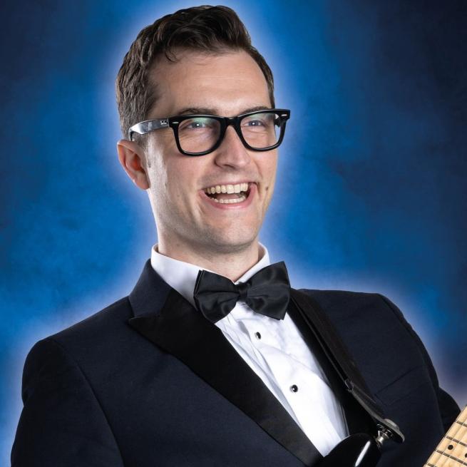 Man in tuxedo and bow tie wearing glasses and smiling against a blue background playing a Fender electric guitar.