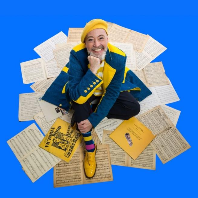 A man with a yellow beret and matching shoes sits smiling on pages of sheet music and Victoria Wood records. 