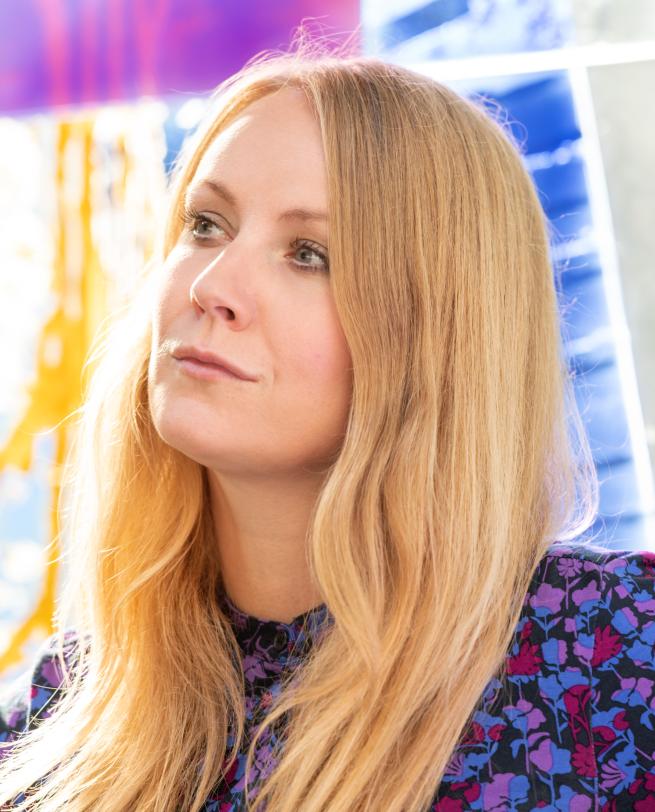 Close-up portrait of a woman with long blonde hair, looking off-camera with a thoughtful expression, wearing a purple and blue floral patterned blouse.