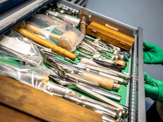 Inside a museum collections store, gloved hands open a drawer full of old medical instruments.