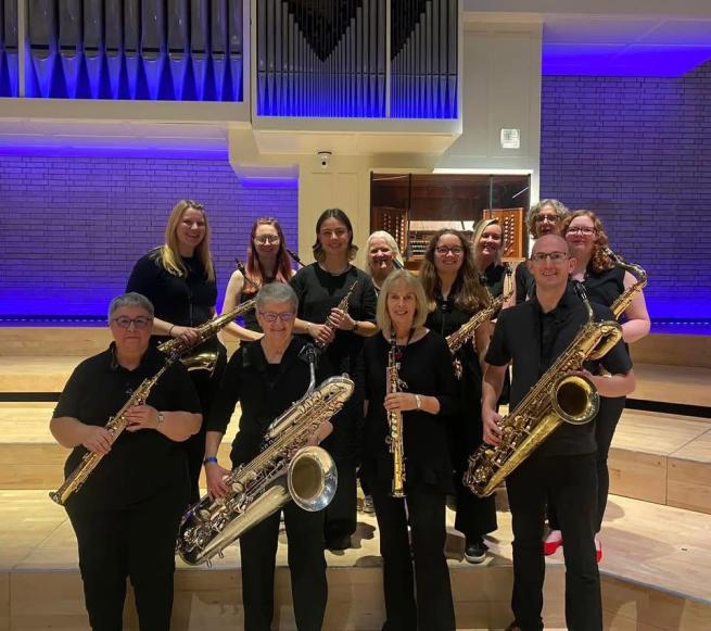 A group of around a dozen people all dressed rather smartly in black outfits and holding an impressive array of saxomophones of varying sizes, definitely one for the saxophone enthusiast