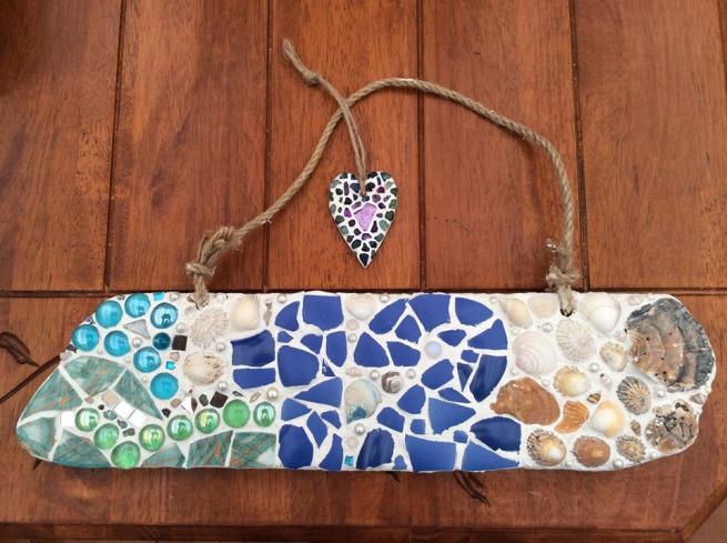 Image shows a mosaic piece on driftwood with shells and tiles embedded.