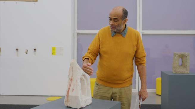 A mean wearing a yellow jumper touching a small stone sculpture with the back of his hand.