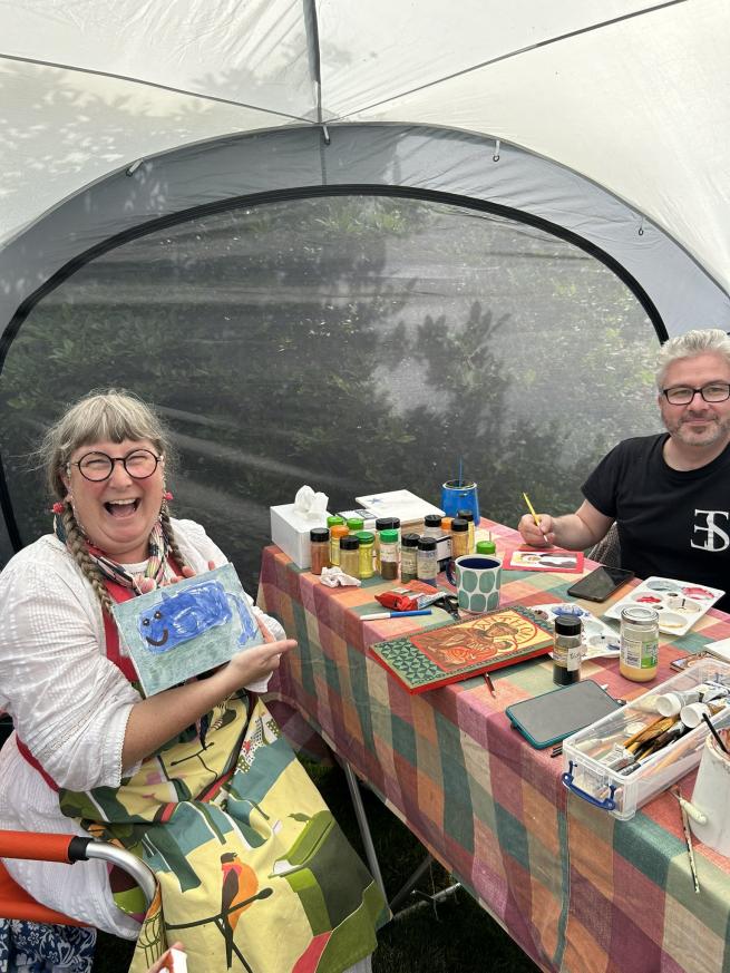 2 artists are sat at a table under a gazebo, the lady who is sat to the right holds up a painting of a whale and is laughing
