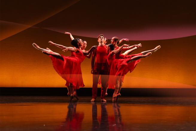 A male dancer in red attire stands in the centre surrounded by 5 female dancers. They surround the central image and pose, balancing on one foot with the other leg elevated to the back.