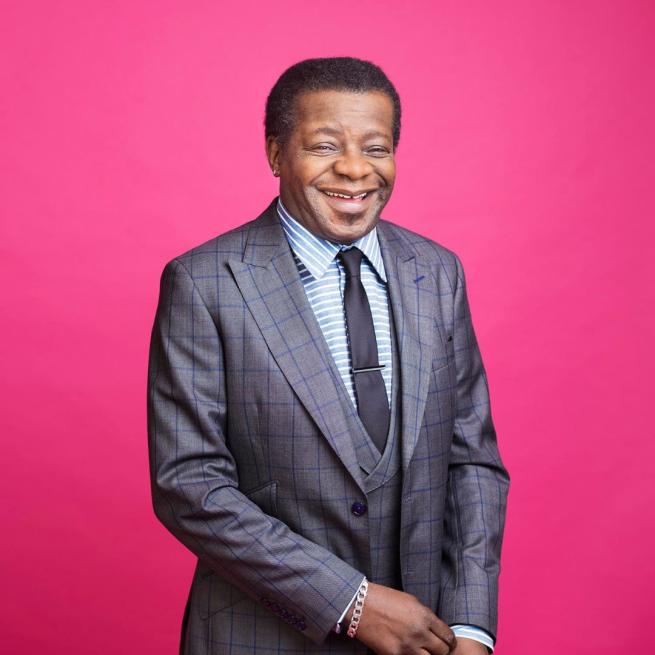 Wearing a grey suit in front of a pink background, Stephen K Amos stands laughing.