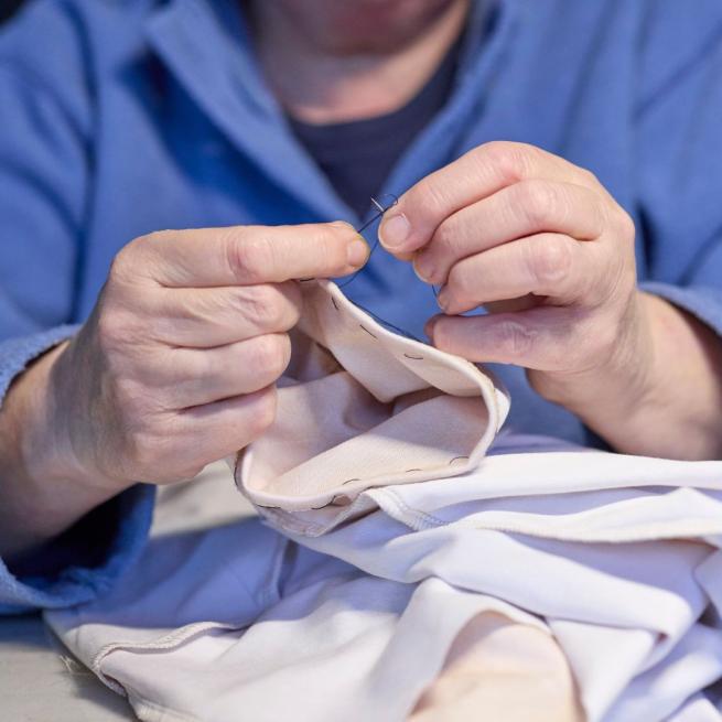 A photograph of a person threading a needle whilst holding a garment which has the hem folded and tacked with black stitches