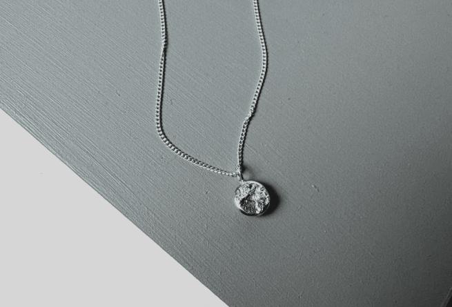 Image shows a textured ingot necklace on top of a grey tabletop. 
