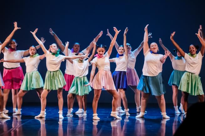 A group of dancers on stage with their arms stretches out in all directions, dressed in white t-shirts and various coloured skirts