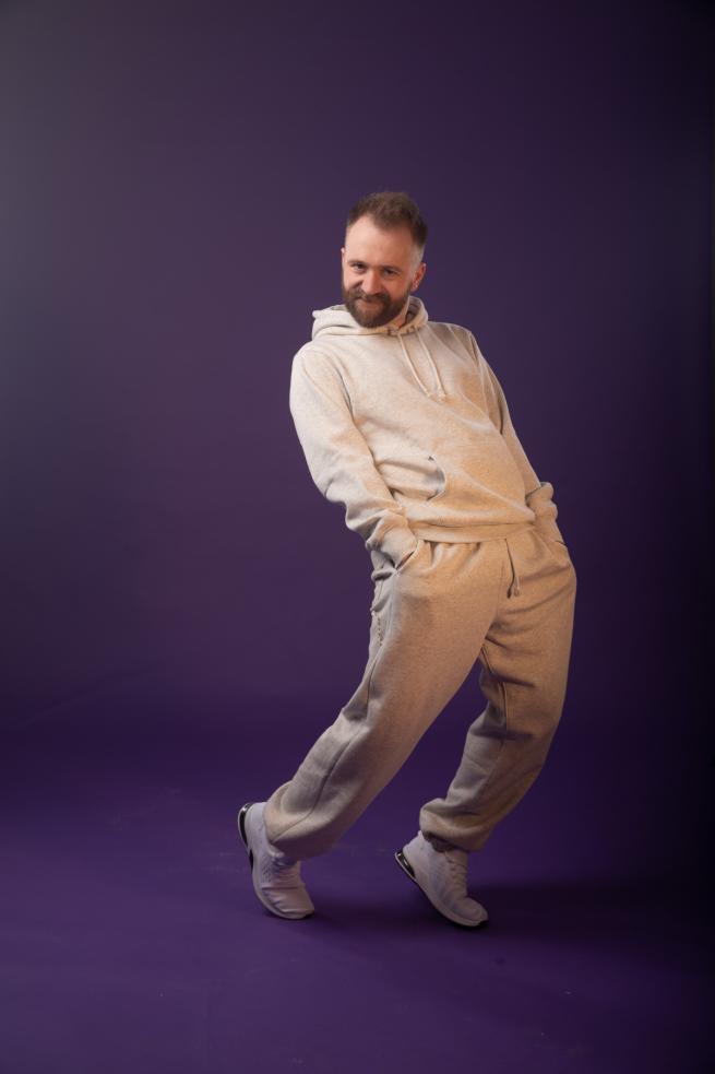 A white man with a grey tracksuit poses cheekily against a purple background.