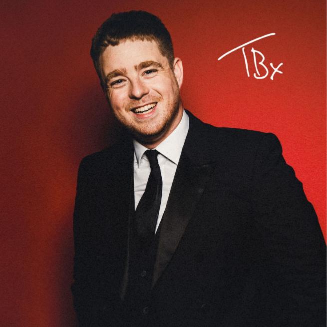 Tom Ball in a suit smiling, with hands casually in his pockets against a red backdrop.
