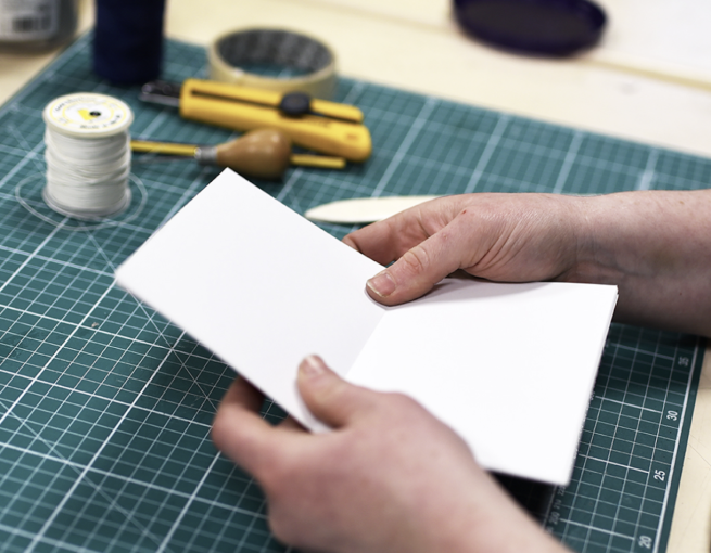 A pair of hands folding paper to make a book, surrounded by bookbinding tools