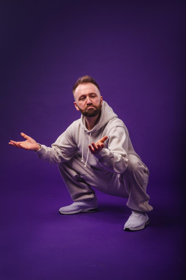 A white man with a beard wearing a full white tracksuit, squatting and frowning warm but goofy manner on a purple background