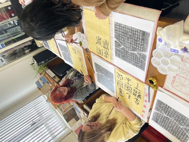 Three people gathered around a table - painting calligraphy on large yellowed paper
