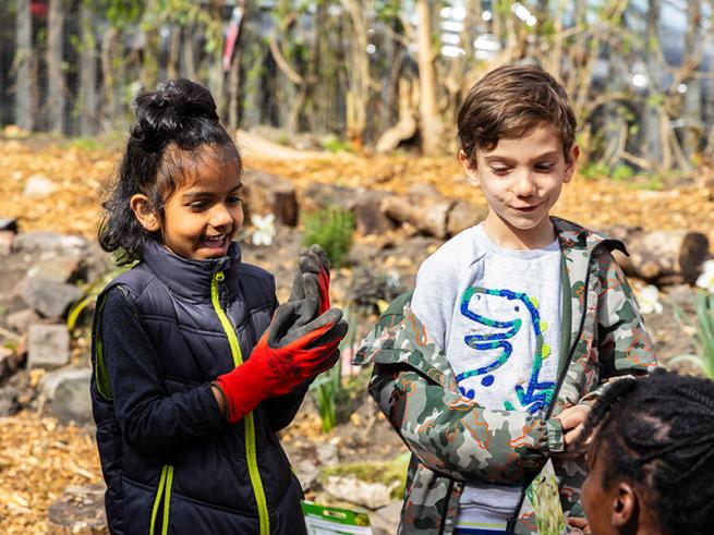 Two children smile and engage in a playful outdoor setting. One wears red gardening gloves, holding soil. The backdrop includes wood chips and plants, evoking adventure and joy.