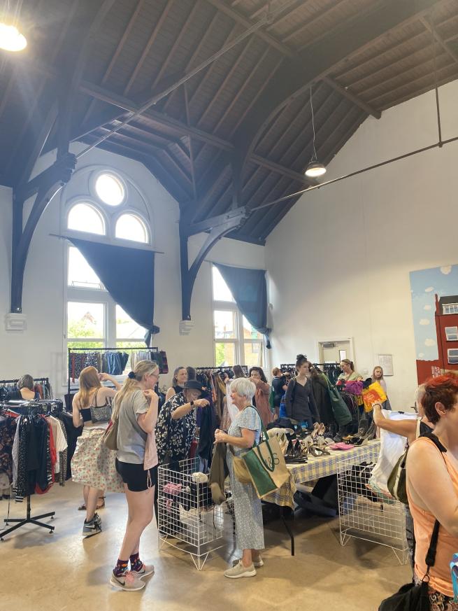 Groups of people browsing rails and holding up items of clothing in a brightly lit room filled with rails and baskets of clothing.