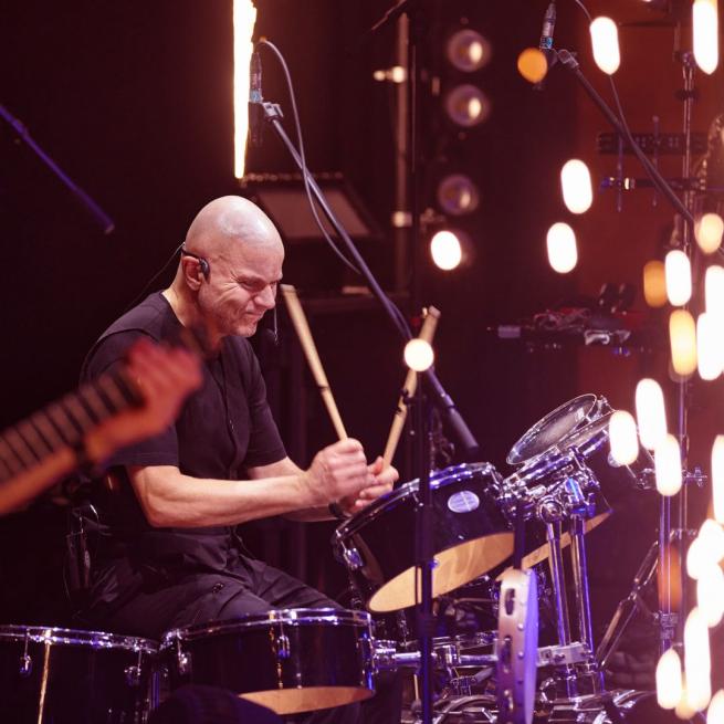 A man plays the drums on stage as sparks fly in the foreground.