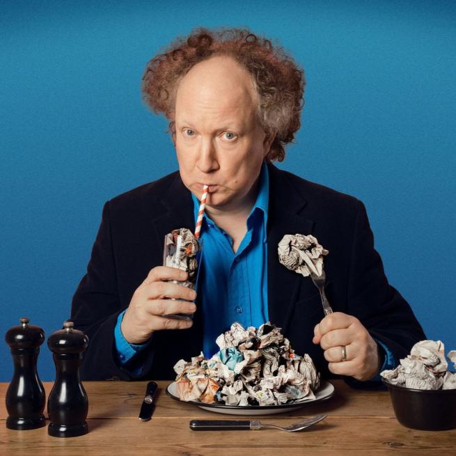 Andy Zaltzman sits at a wooden table, sipping from a glass filled with scrunched up newspaper. In front of him there is a plate, cutlery and bowl also filled with discarded newspaper.