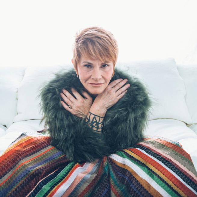 Shawn Colvin smirks with hands across her chest, wearing a furry green jacket, sat on a bright white bed.
