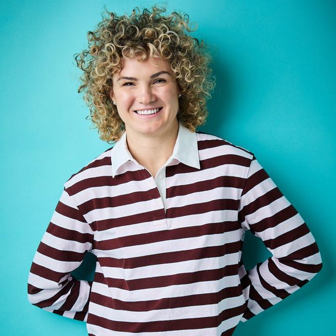 Ellie Kildunne smiles at the camera, wearing a stripy, long-sleeve collared shirt, with hands behind her back, in front of a blue backdrop.