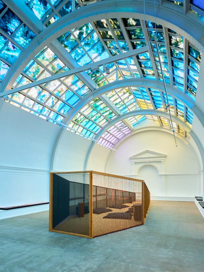 A large white room with a colourful lit ceiling above a wooden coffin shaped oversized sculpture.