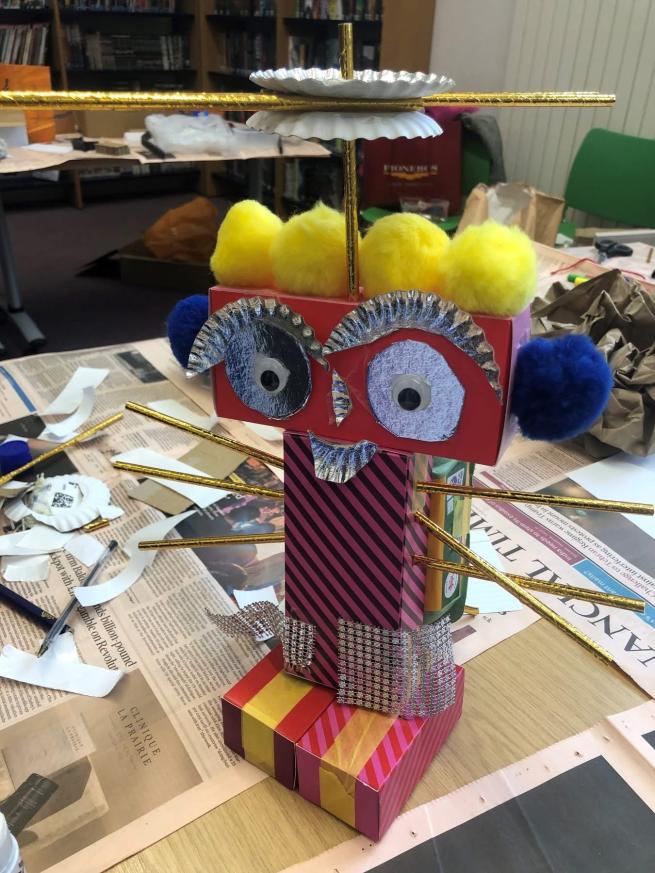 A small 'robot' made from junk materials with yellow pom pom hair, gold straw arms, shiny tin foil eyes and eye brows and stripey body.