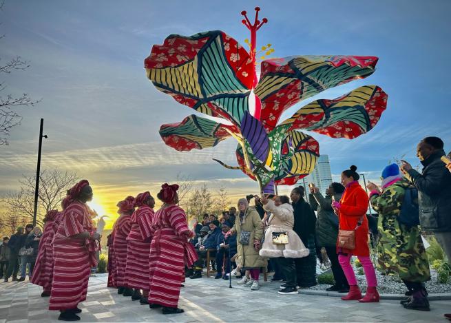 Performers from Yoruba Heritage Group Yorkshire, dressed in red and white striped traditional costumes, dance beside Hibiscus Rising, Yinka Shonibare’s large flower sculpture covered in a colourful Ankara fabric design, in Aire Park, Leeds. Members of the public gather around, watching and taking photographs against a dramatic sunset sky.